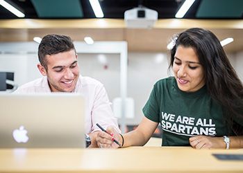 Two MSU students sitting at a table. One of the students is wearing a shirt that says, 