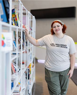 MJ Monteiro stands next to a colorful bookshelf in the Broad Art Museum.