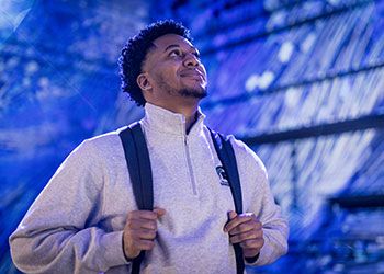 Joshua Buggs wearing a backpack, standing inside a campus building, looking up and smiling.