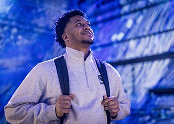 Joshua Buggs wearing a backpack, standing inside a campus building, looking up and smiling.