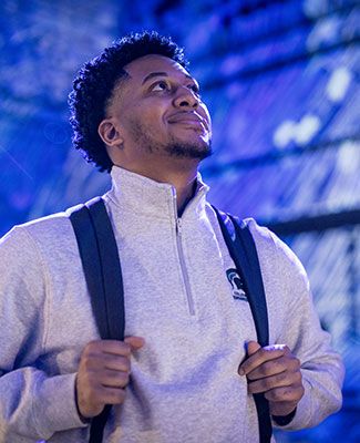 Joshua Buggs, wearing a backpack, standing inside a campus building, looking up and smiling.