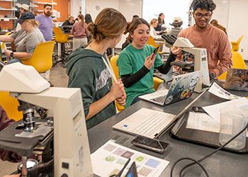 Three students talk around a lab bench in a crowded classroom, with their laptops and microscope nearby.