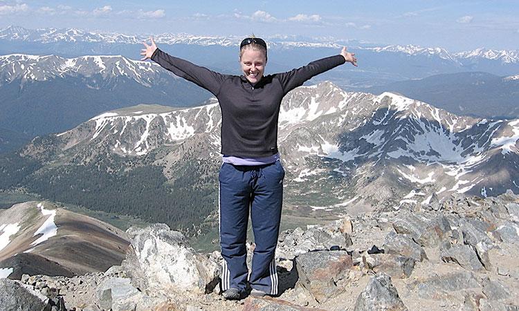 photo of Renee Cherry standing on a mountaintop with other mountains in the background. She is holding her arms out wide.